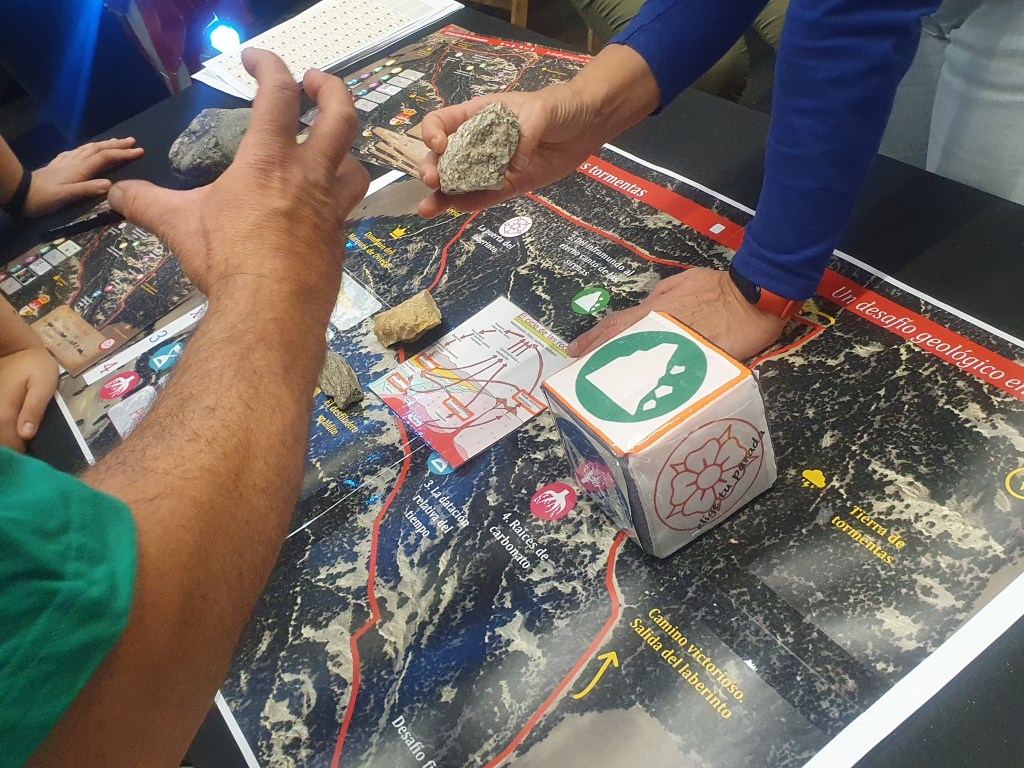 Fotografía en detalle de una de las paradas del juego, donde se ve el dado y las rocas implicadas en la explicación, y uno de los jugadores mostrando gran interés por aprender jugando.