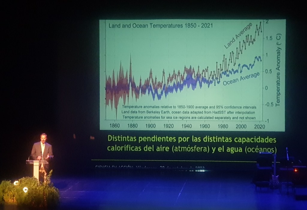 Fotografía de Miguel Viñas durante su conferencia de clausura. Se puede ver una de las diapositivas de su presentación donde compara la temperatura del aire y del océano en el tiempo ciendo como va en aumento y más se estan distanciando la una de la otra.
