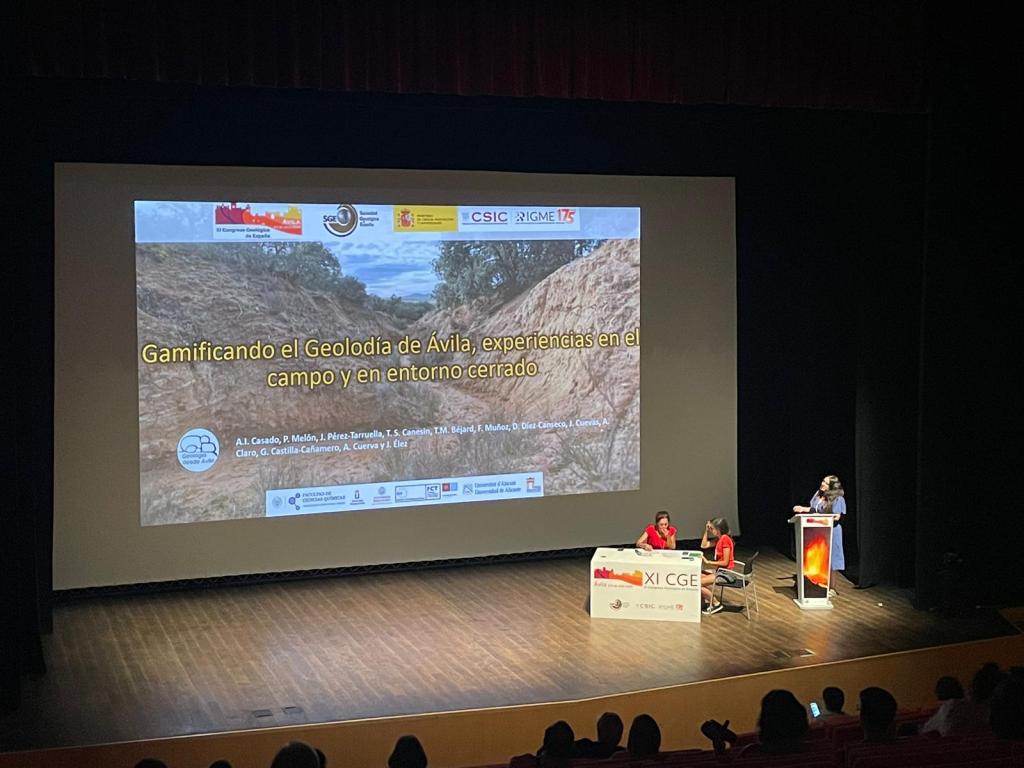 Nuestra compañera @anabelgeoraman durante la exposición del trabajo en la Sesión de Didáctica y divulgación del Congreso Geológico de España el pasado 3 de julio. Se puede ver a Ana Isabel detrás del atril, señalando a la pantalla con la proyección de la presentación de la ponencia, donde se puede leer "Gamificando el Geolodía de Ávila, experiencias en el campo y en entorno cerrado". También se ve a las dos geólogas responsables de la sesión sentadas alrededor de la mesa de organizadoras.