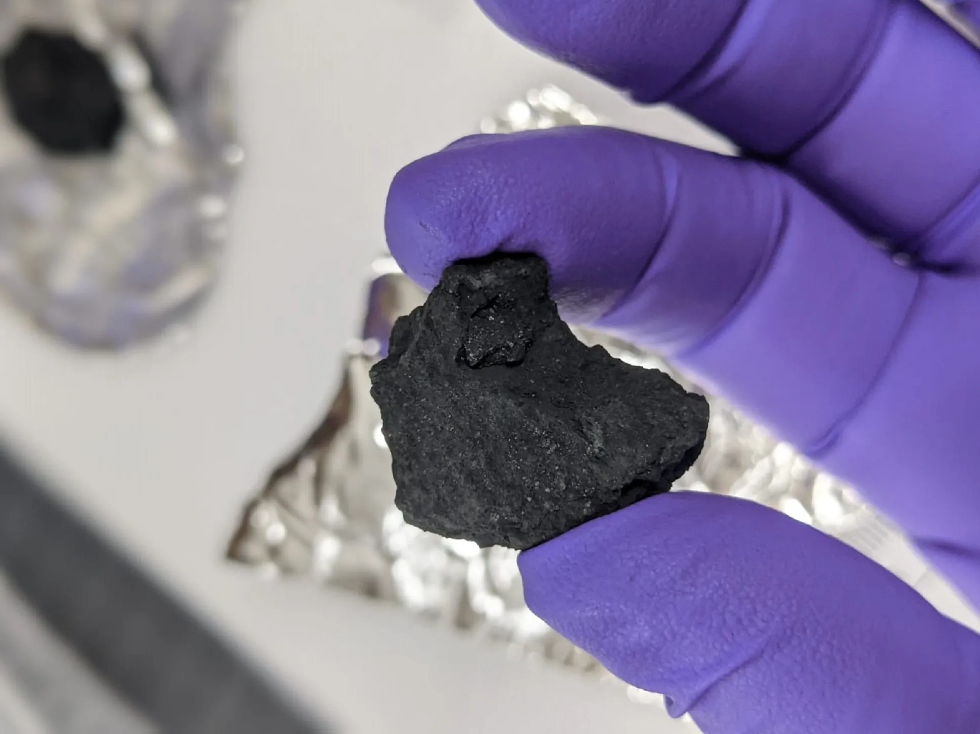 Figura 11. Primer plano de un fragmento del meteorito Winchcombe sostenido entre los dedos índice y pulgar de una mano enguantada con guantes de látex morado. El fragmento es de color negro mate, con superficie rugosa e irregular. Al fondo, desenfocado, se observa papel de aluminio y una superficie de laboratorio. La imagen fue tomada durante los análisis científicos realizados para estudiar su contenido en agua. Este meteorito, una condrita carbonácea, fue recuperado pocas horas después de su caída gracias a la colaboración ciudadana, lo que permitió preservar su composición original. Fuente: Museo de Historia Natural, Londres.