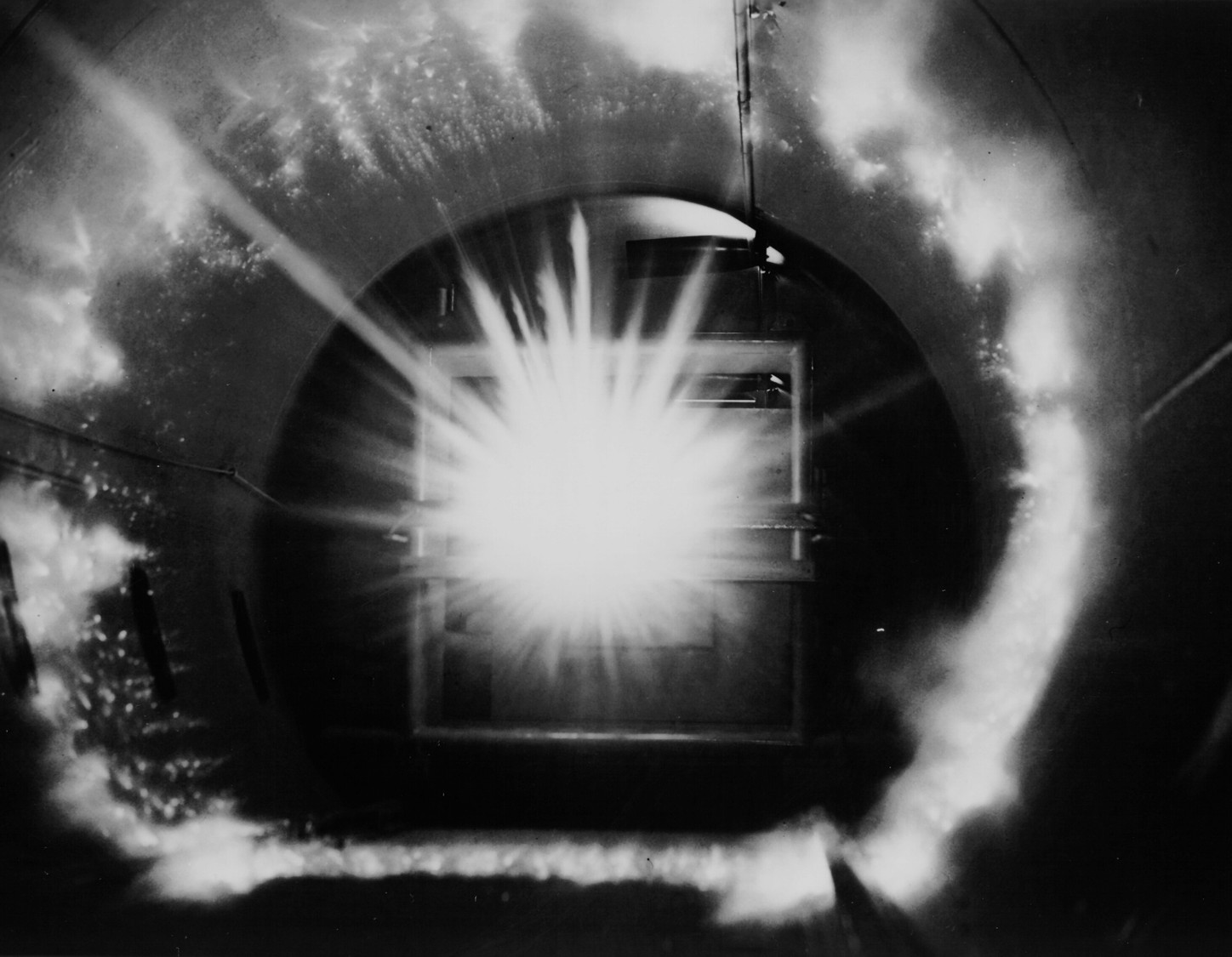 Fotografía en blanco y negro de un experimento de impacto a hipervelocidad realizado en laboratorio. En el centro de la imagen se observa un fogonazo brillante, con rayos de luz que se expanden radialmente hacia los bordes de una cámara circular metálica. El destello ilumina las paredes internas del túnel, revelando una estructura cilíndrica. El fenómeno simula el impacto de partículas a velocidades extremas, como las que ocurren en el espacio. Fuente: NASA / Ames Research Center, Mountain View, California.