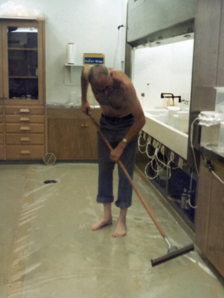 La imagen en color muestra a un hombre mayor, descalzo, sin camisa y con los pantalones remangados hasta media pantorrilla, limpiando el suelo de un laboratorio con una mopa o escurridor de goma. Se trata del científico Clair Patterson, reconocido por su trabajo sobre la datación de la Tierra y por alertar sobre la contaminación por plomo.

El laboratorio tiene un aspecto ordenado, con muebles de madera clara, una campana de extracción a la derecha, y varias tuberías y cables visibles bajo la encimera. Encima de un dispensador de papel, hay una caja azul y amarilla con la etiqueta "Saran Wrap" (una marca de film plástico). En el suelo parece haber una película plástica transparente que el científico está limpiando cuidadosamente.

Esta escena refleja el nivel extremo de limpieza que Patterson mantenía en su laboratorio para evitar cualquier mínima contaminación externa, especialmente de plomo, ya que su trabajo requería mediciones ultrasensibles. Gracias a estas medidas, fue pionero en establecer uno de los primeros laboratorios de ambiente limpio (clean room) en el mundo.
