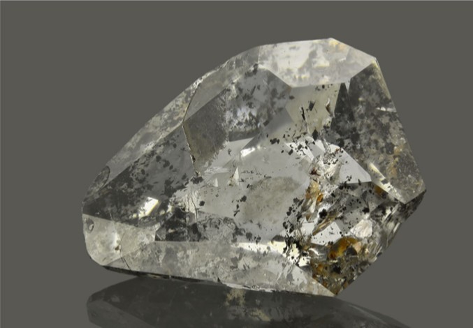 Figura 5. Fotografía de un diamante transparente y facetado sobre un fondo gris neutro. En su interior se observan numerosas inclusiones minerales, visibles como manchas oscuras e irregulares, que corresponden a ringwoodita y circón. El diamante tiene forma asimétrica, con múltiples caras planas y bordes definidos. Procede de la República Centroafricana. En Lorenzon et al. (2022).