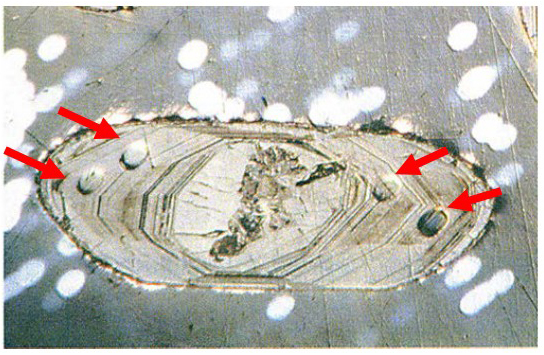 La imagen muestra una fotografía microscópica a color de un cristal de circón, un mineral muy resistente utilizado frecuentemente para la datación radiométrica de rocas antiguas. El circón aparece como una forma ovalada y alargada, con una serie de líneas concéntricas internas que marcan su crecimiento por capas, similares a los anillos de un árbol. En el centro del cristal se observa una zona oscura e irregular, posiblemente una inclusión o una zona alterada.

A lo largo del borde del circón se pueden ver cuatro pequeños orificios circulares, señalados con flechas rojas. Estos agujeros fueron hechos de forma controlada mediante el impacto de haces de iones que vaporizaron mínimas cantidades del mineral para permitir la medición de la relación entre uranio y plomo en puntos específicos del cristal. Esta técnica permite determinar con gran precisión la edad del circón.

Este circón procede de una roca muy antigua llamada gneis de Acasta, ubicada en Canadá. Su datación ha revelado una edad aproximada de 4.000 millones de años, lo que lo convierte en uno de los minerales más antiguos encontrados en la Tierra.

Fuente de la imagen: Adaptado de York (1993).