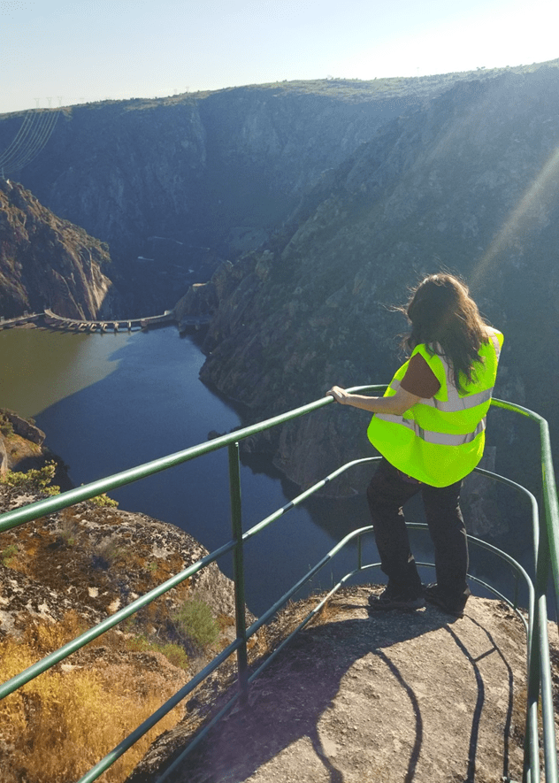 Ana Isabel con chaleco reflectante amarillo fosforito y camiseta roja observa el paisaje desde un mirador rocoso con barandilla metálica (el mirador de Felipe), en lo alto de un profundo cañón del parque natural de los Arribes del Duero. Frente a ella, al fondo del barranco, fluye el río Duero encajado entre paredes verticales de roca. Se distingue una presa hidroeléctrica con compuertas y un conjunto de cables de alta tensión que atraviesan la garganta. El paisaje combina vegetación dispersa en las laderas, formaciones rocosas y una vista impresionante del valle. La luz del sol, en ángulo, proyecta sombras alargadas y realza los contrastes del terreno.