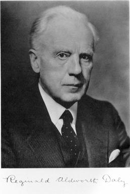  Retrato en blanco y negro de Reginald Aldworth Daly, mayor con el pelo claro peinado hacia atrás, vestido con traje oscuro, camisa blanca y corbata; aparece de medio cuerpo, mirando a la cámara con expresión seria, y bajo la fotografía se ve una firma manuscrita.