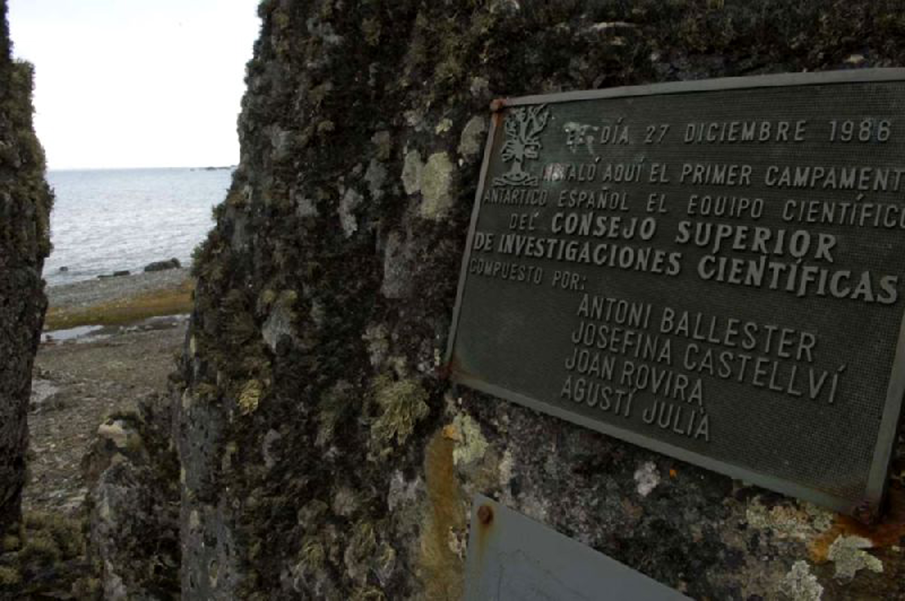 Qué se ve en general
Fotografía en color tomada en exterior, en un paisaje costero frío. La imagen está dominada por una roca grande y oscura, cubierta de líquenes en tonos verdosos y blanquecinos. A la izquierda, entre dos paredes de roca, se abre un pasillo estrecho que deja ver el mar y una franja de costa pedregosa.

Elemento principal: la placa conmemorativa
En la parte derecha de la imagen, fijada a la roca, hay una placa metálica rectangular de color oscuro, con letras en relieve de color claro. La placa está colocada en ligera diagonal, como inclinada hacia la cámara. En la esquina superior izquierda aparece un pequeño emblema grabado.

Texto legible en la placa
El texto, en mayúsculas, indica una fecha y conmemora el primer campamento español. Se lee:

“DIA 27 DICIEMBRE 1986”

“INSTALÓ AQUÍ EL PRIMER CAMPAMENTO ANTÁRTICO ESPAÑOL”

“EL EQUIPO CIENTÍFICO DEL CONSEJO SUPERIOR DE INVESTIGACIONES CIENTÍFICAS”

“COMPUESTO POR:”

“ANTONI BALLESTER”

“JOSEFINA CASTELLVÍ”

“JOAN ROVIRA”

“AGUSTÍ JULIÀ”

Entorno y sensación de lugar
La combinación de roca húmeda con líquenes, costa pedregosa y mar al fondo refuerza la idea de un punto de desembarco o campamento en un entorno remoto.