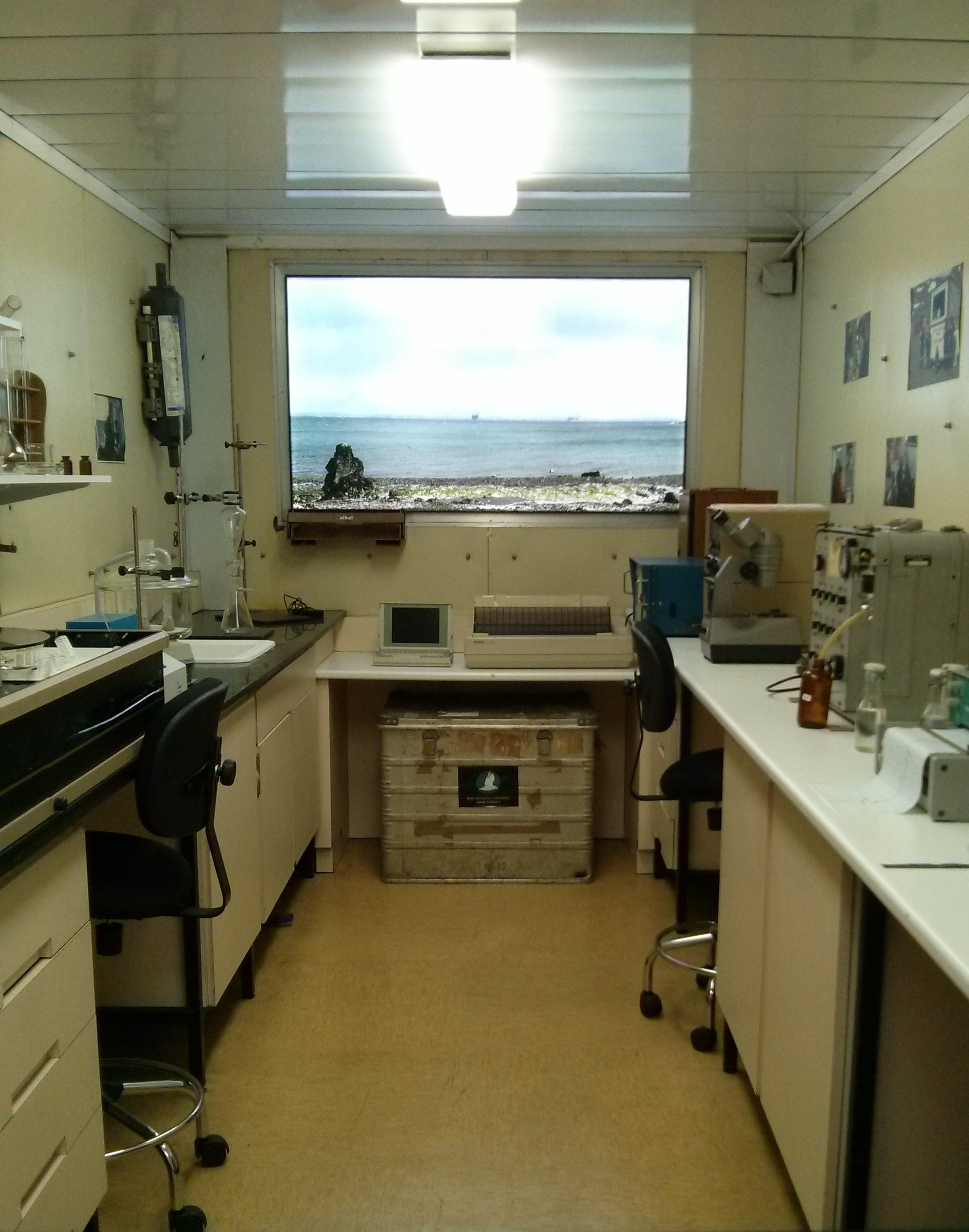 Qué se ve en general
Fotografía en color de un laboratorio pequeño y estrecho, visto desde la entrada como un pasillo. La cámara está centrada, mostrando el espacio en perspectiva hacia el fondo. El laboratorio está bien iluminado por una luz rectangular en el techo.

Distribución del espacio
Hay mesas de trabajo a ambos lados: una a la izquierda y otra a la derecha, con armarios y cajones debajo. En cada lado hay sillas giratorias negras con ruedas, colocadas junto a las mesas. El suelo es liso, de color beige.

Ventana y vista exterior
Al fondo, ocupando casi toda la pared, hay una ventana grande. A través de ella se ve el mar en calma, una franja de costa pedregosa y un cielo claro con nubes suaves. Cerca de la orilla se distingue una roca oscura prominente.

Material y equipos (lado izquierdo)
Sobre la mesa izquierda hay material de vidrio de laboratorio: recipientes transparentes, tubos y soportes metálicos con pinzas. También se ve una estantería pequeña con más utensilios y frascos. La pared tiene algunos papeles o fotos pequeñas pegadas.

Material y equipos (lado derecho)
En la mesa derecha hay equipos de laboratorio más voluminosos, de aspecto instrumental, con botones o mandos. También aparece un frasco pequeño ámbar (marrón) y otros recipientes. En la pared derecha hay varias fotografías o láminas colocadas en fila.

Zona central del fondo
Debajo de la ventana hay una mesa o repisa con un monitor pequeño y un equipo rectangular. En el suelo, centrada, hay una caja metálica grande con aspecto de contenedor de transporte, con una pegatina o emblema en el frontal.

Impresión general
La imagen muestra un laboratorio funcional, compacto y ordenado, con la particularidad de la gran ventana con vista directa al mar, que sitúa el trabajo científico en un entorno polar costero.