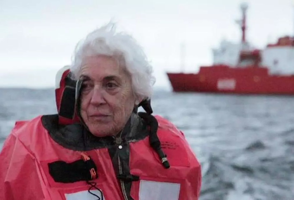 Qué se ve en general
Fotografía en color, tipo fotograma de documental, tomada en un entorno marino frío. El fondo muestra agua gris con oleaje suave y un cielo pálido.

Persona y encuadre
En primer plano aparece Pepita Castellví (Josefina Castellví), una mujer mayor, encuadrada de pecho hacia arriba. Tiene el pelo blanco, corto y con volumen, algo despeinado por el viento. Su rostro está girado ligeramente hacia la izquierda y su expresión es serena y pensativa, como si estuviera observando algo fuera de plano.

Ropa y equipo
Lleva una chaqueta gruesa de color rojo intenso con detalles negros y cierres visibles. En la parte frontal se ven correas, costuras y un gran panel claro rectangular, como un parche reflectante o identificativo. El cuello está subido y acolchado, indicando protección contra el frío y el viento.

Fondo: barco
A la derecha, en segundo plano y desenfocado, se distingue un barco grande rojo sobre el mar. El barco aparece algo borroso por la profundidad de campo, pero su color y forma destacan claramente.

Impresión general
La escena transmite un momento de viaje y despedida en un entorno antártico: Pepita en primer plano, abrigada, con el mar y un barco de apoyo al fondo.