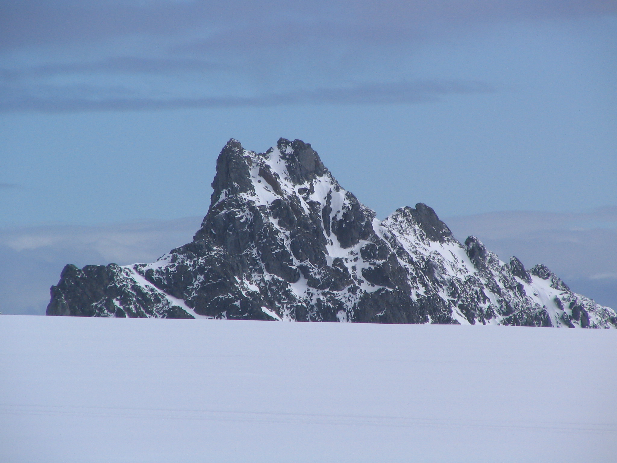 Qué se ve en general
Fotografía en color de un paisaje polar abierto. En primer plano domina una gran extensión de nieve lisa, casi uniforme, que ocupa aproximadamente la mitad inferior de la imagen.

Elemento principal: el pico
En el centro se eleva un macizo rocoso oscuro con forma de pico. La roca es casi negra o gris muy oscuro y está marcada por manchas y franjas de nieve que se acumulan en grietas y laderas, creando un contraste fuerte entre blanco y oscuro. La cumbre es irregular y afilada, con varios salientes, y a la derecha se prolonga en una cresta más baja.

Fondo: cielo y atmósfera
El cielo ocupa la mitad superior de la imagen y es de color azul pálido, con bandas finas de nubes horizontales. No se ven personas, edificios ni vegetación.

Impresión general
La escena transmite aislamiento y amplitud: un pico rocoso emergiendo sobre una planicie nevada, en un entorno frío y silencioso.